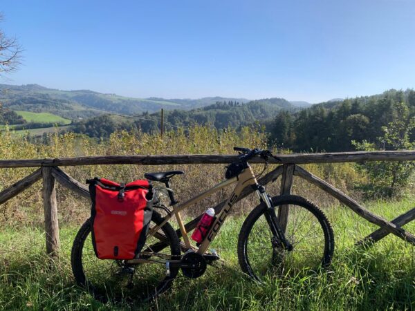 Bicicletta con le borse rosse appoggiata a una staccionata di legno, con le colline dell'Appennino bolognese