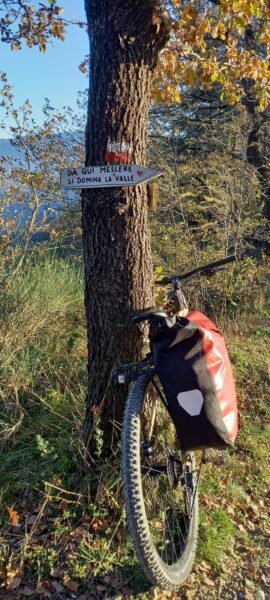 Cartello «Da qui messere si domina la valle» su un tronco segnato di bianco e rosso, con la bicicletta