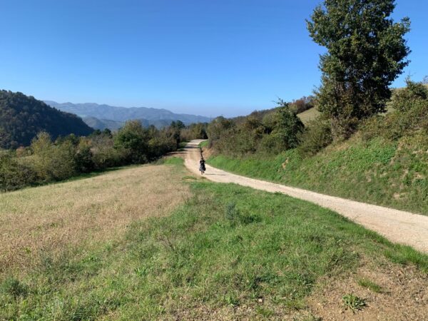 Sterrato di crinale tra prati e cespugli, con un ciclista e le montagne all'orizzonte